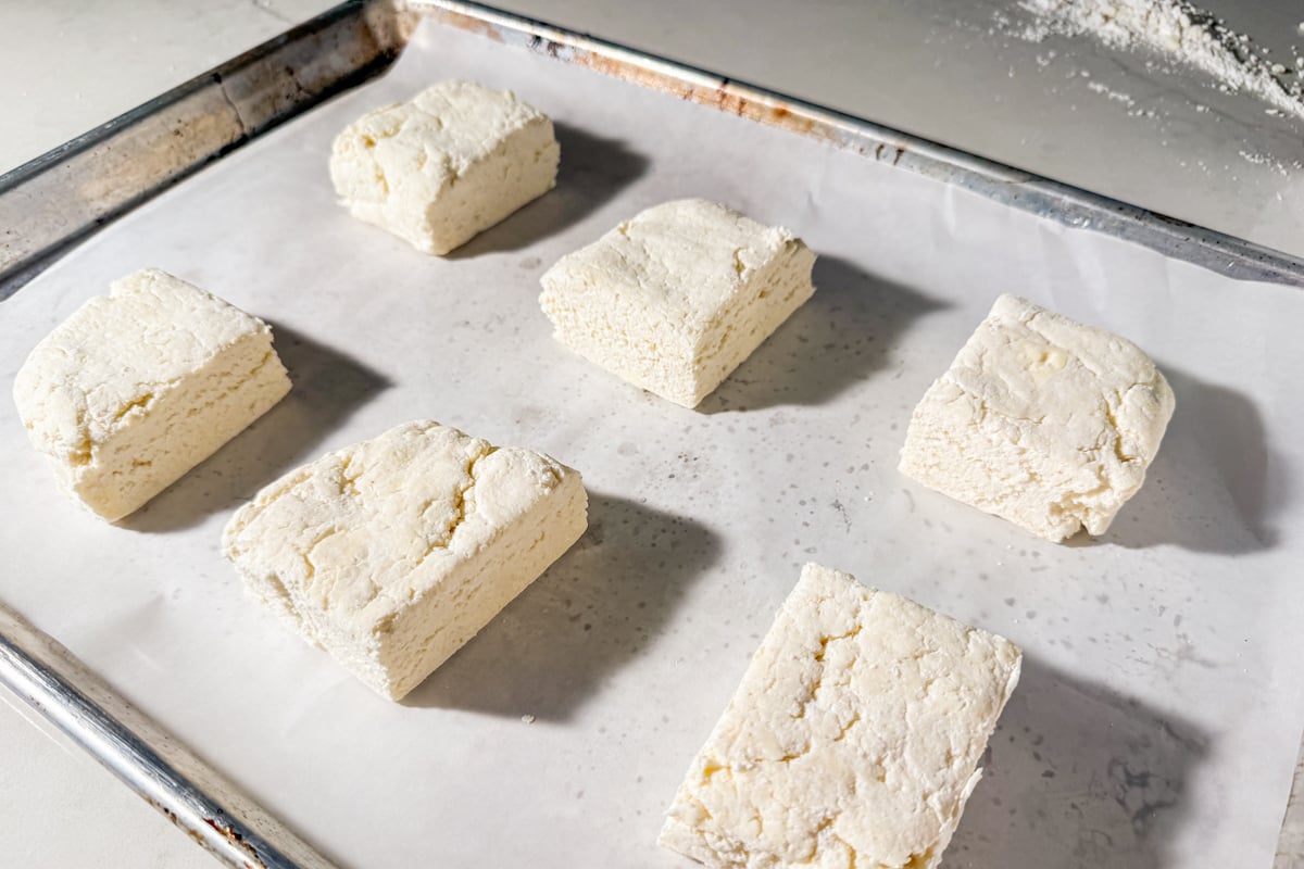 Six unbaked square biscuits made from dough are spaced apart on a parchment-lined baking sheet, ready to be baked. The biscuits have a slightly rough texture and are light in color.