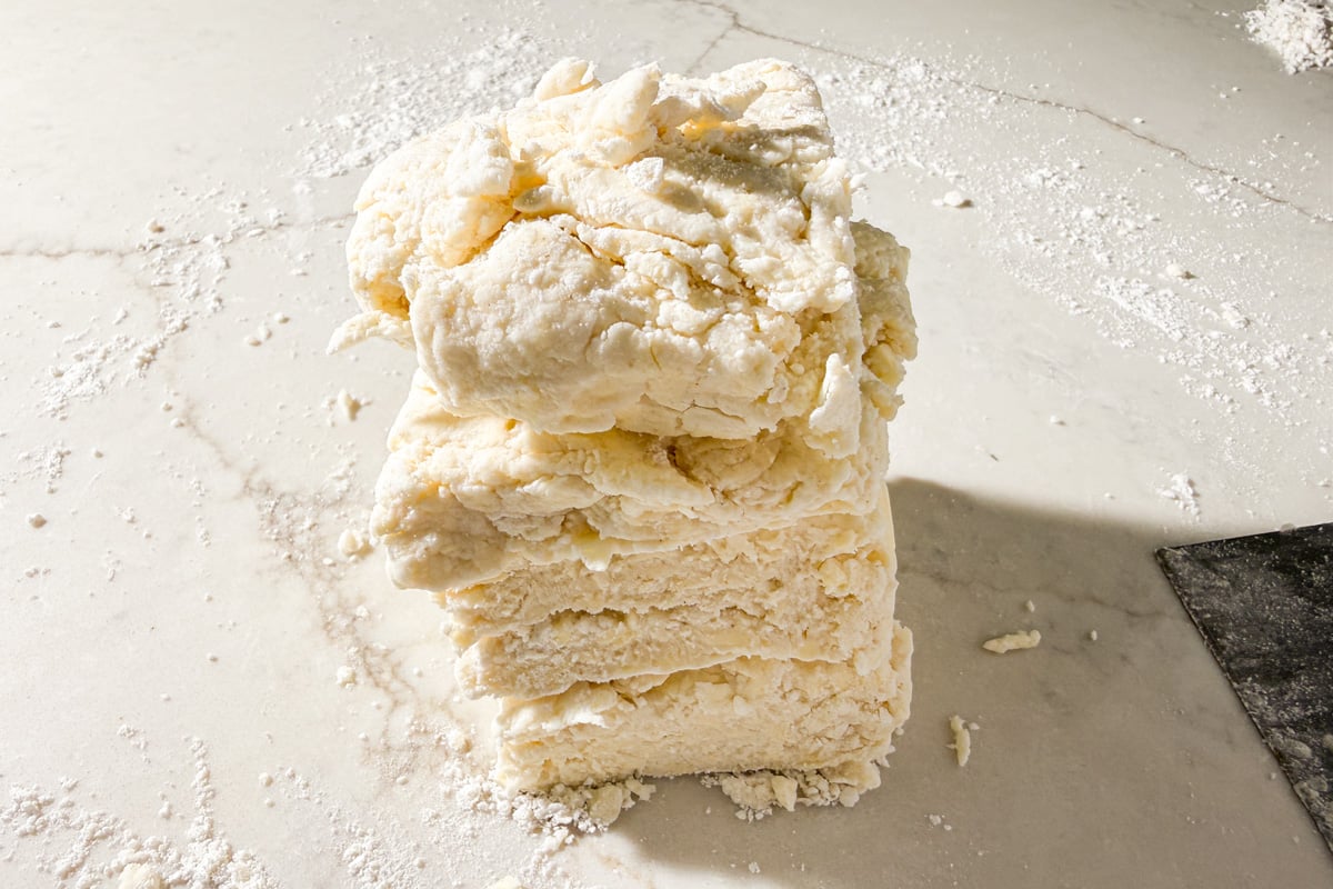 A stack of rough, flaky biscuit dough sits on a flour-dusted marble countertop, ready to be shaped or cut. A metal dough scraper lies nearby.