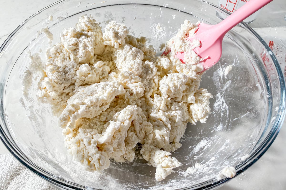 A glass bowl filled with partially mixed dough, some dry flour still visible, and a pink spatula resting in the mixture on a white countertop.