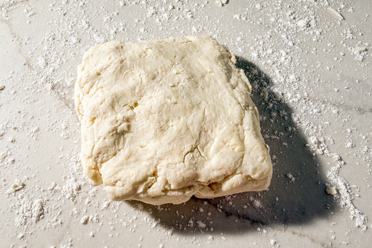 A roughly shaped square of biscuit dough rests on a floured marble surface, with scattered flour around it.