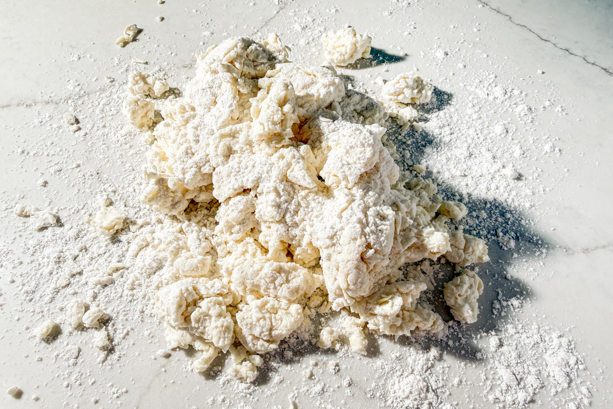 A pile of raw, crumbly dough and loose flour sits on a white, lightly floured marble surface, ready to be kneaded or mixed further.