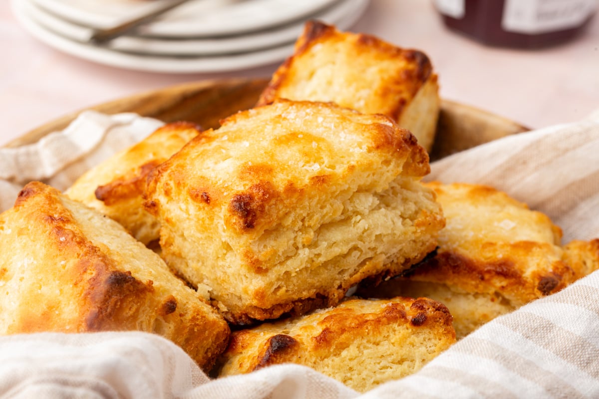 A close-up of golden, flaky biscuits stacked in a basket lined with a beige cloth, with plates visible in the blurred background.