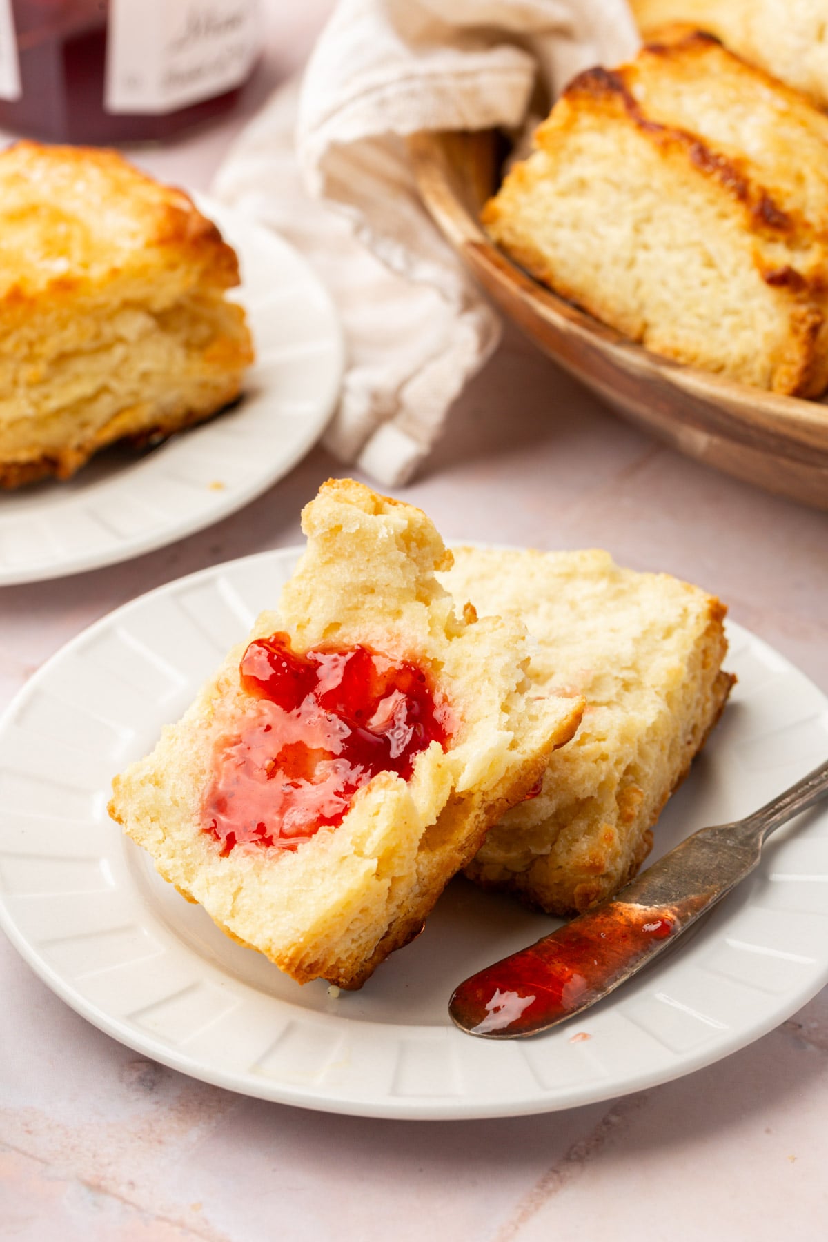 A flaky biscuit split open on a white plate, spread with red strawberry jam. A butter knife with jam rests beside it, and more biscuits are visible in the background.