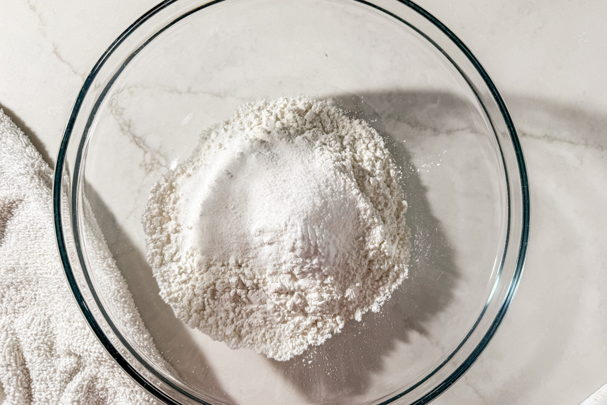 A clear glass bowl containing a mound of white flour and sugar sits on a white marble surface next to a textured white cloth.