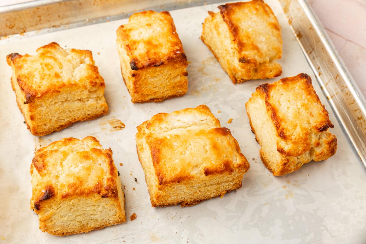 Six golden-brown, flaky biscuits are arranged on a parchment-lined baking sheet. The biscuits have crisp edges and soft, layered interiors, and the tray rests on a light surface.