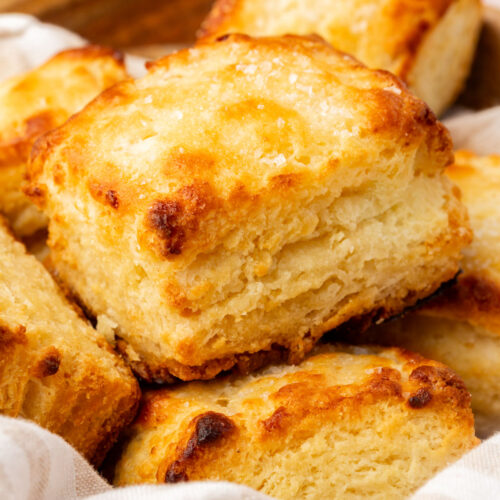 A close-up of golden, flaky homemade biscuits stacked in a basket lined with a white and beige striped cloth.