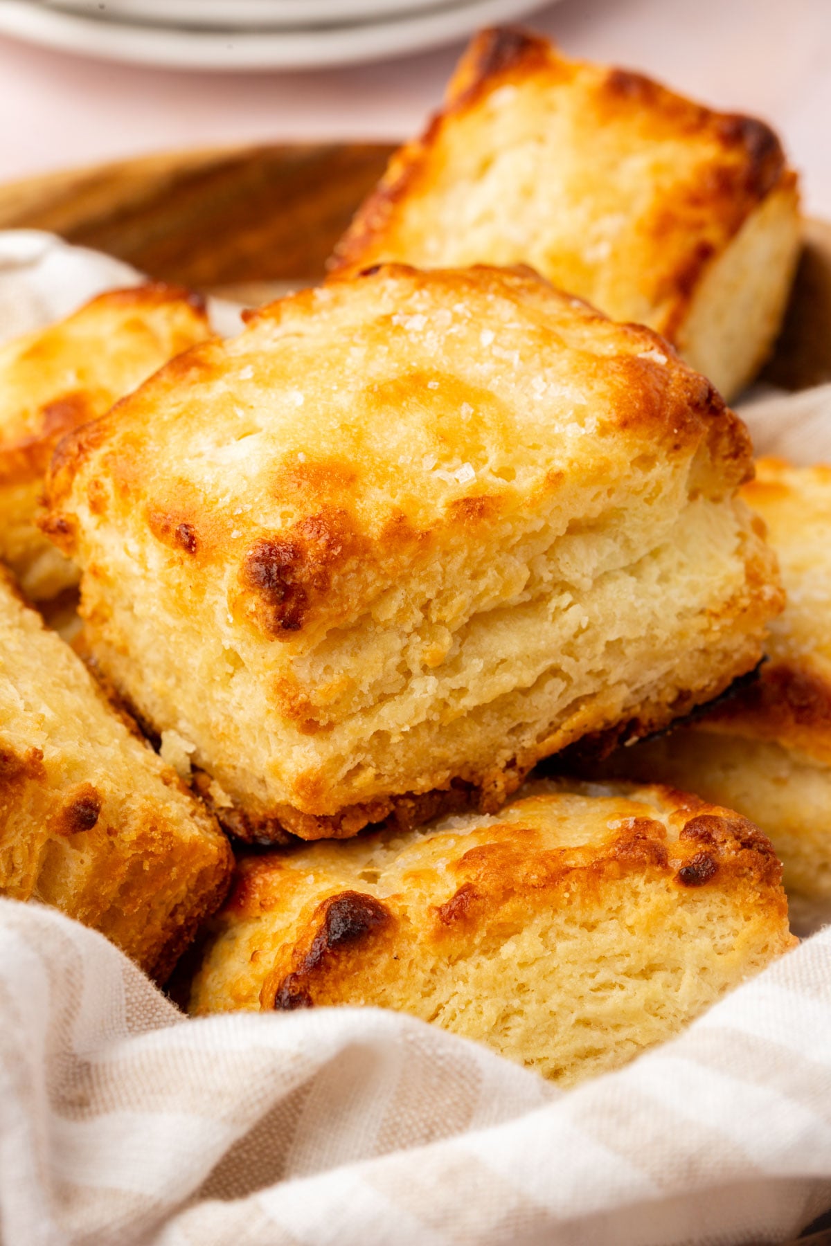 A close-up of golden, flaky homemade biscuits stacked in a basket lined with a white and beige striped cloth.