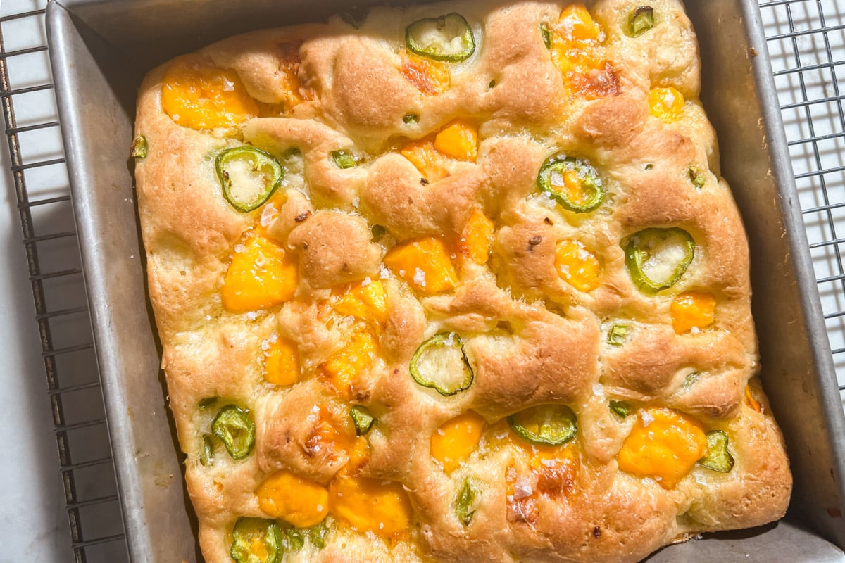 A square pan of golden-brown gluten-free focaccia bread topped with melted cheddar cheese and sliced green jalapeños, cooling on a wire rack.