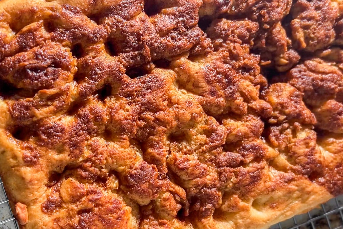 A close-up of a golden-brown, textured cinnamon roll bread, resting on a cooling rack in bright natural light.