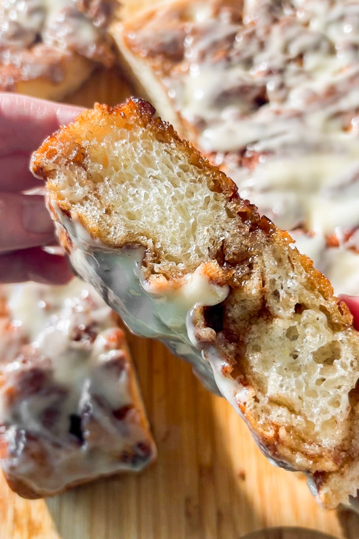 A close-up of a hand holding a gooey cinnamon roll focaccia slice topped with melted cream cheese icing, with more cinnamon roll focaccia slices and icing visible in the background on a wooden surface.