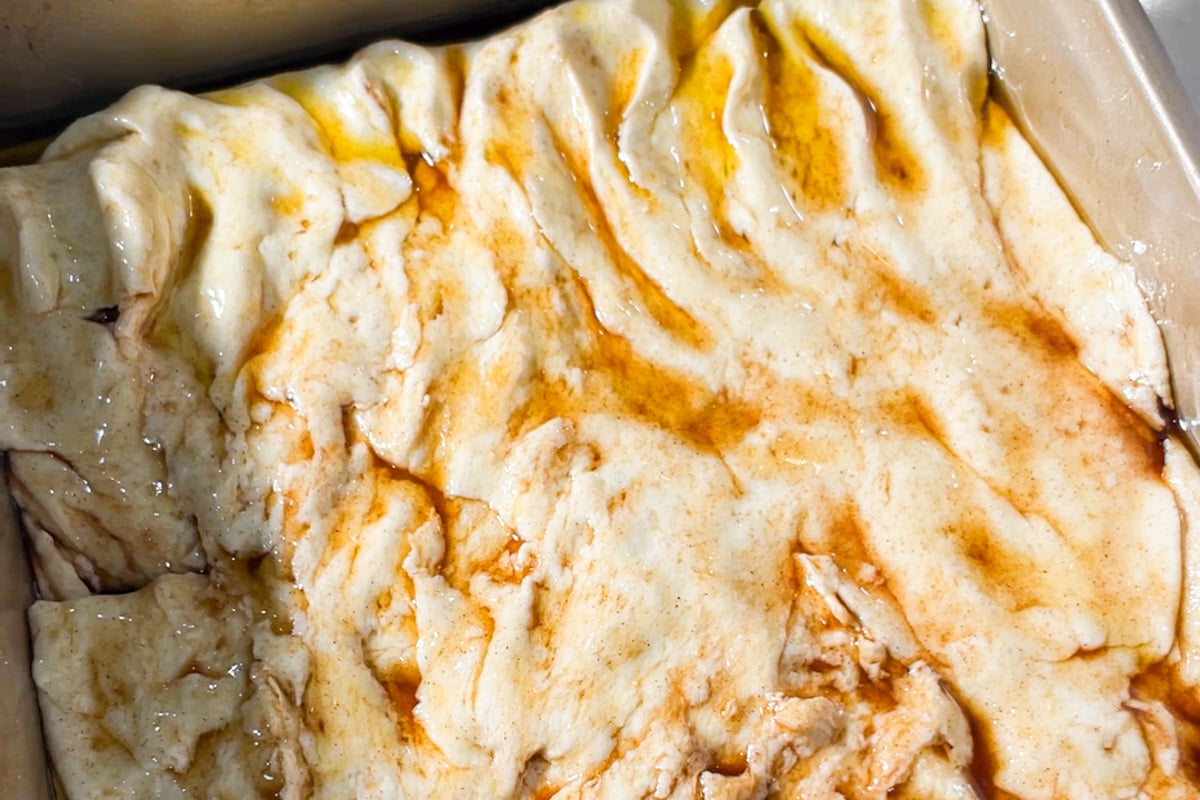 Close-up of unbaked cinnamon roll focaccia dough in a baking dish, drizzled with melted butter and cinnamon sugar, ready for proofing.