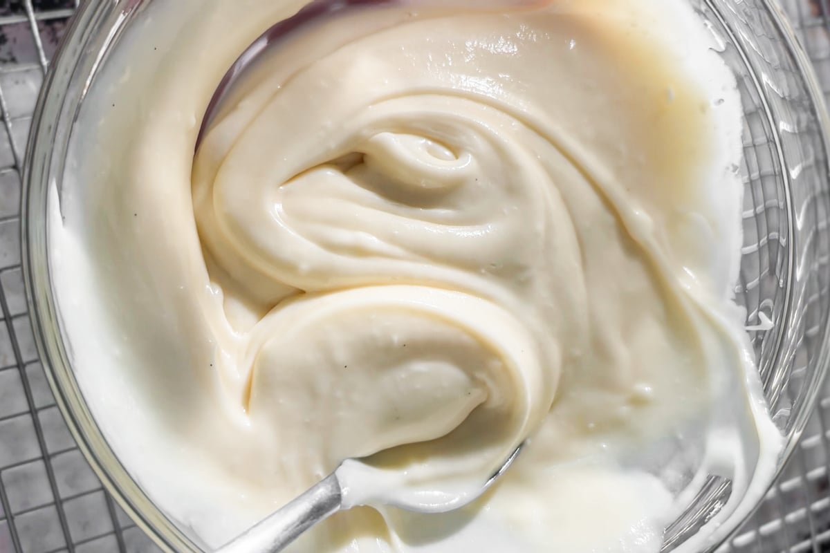 A close-up of a glass bowl filled with smooth, cream cheese frosting, with a spoon partially visible, sitting on a wire rack.