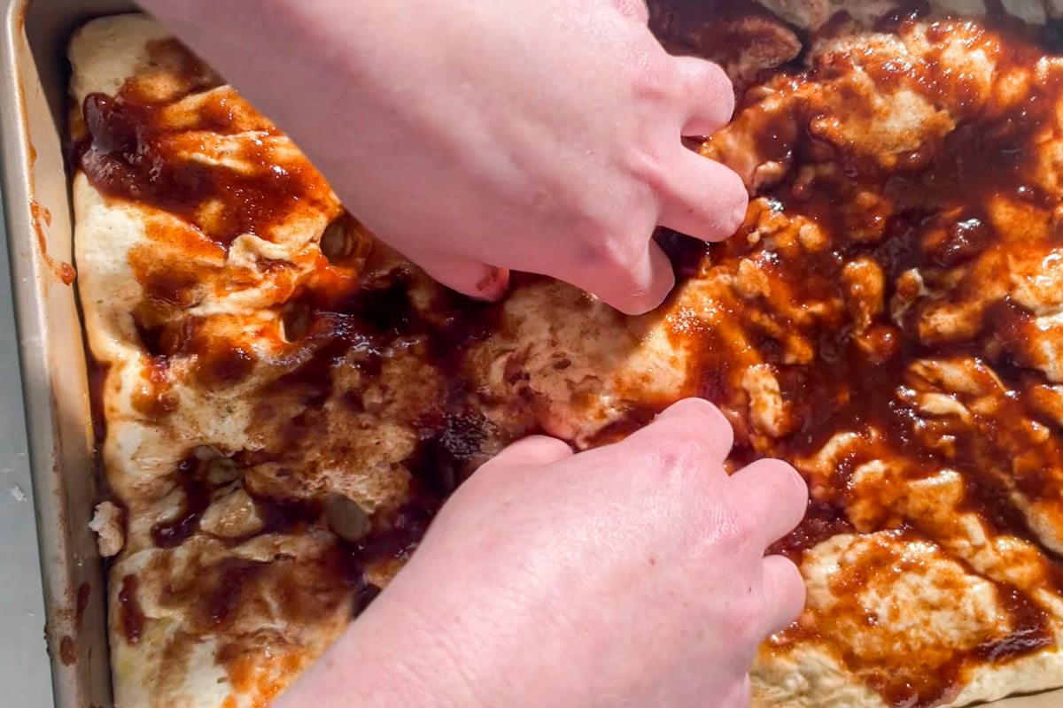 Two hands are dimpling gluten-free cinnamon roll focaccia dough covered with a layer of cinnamon butter mixture in a rectangular baking pan.