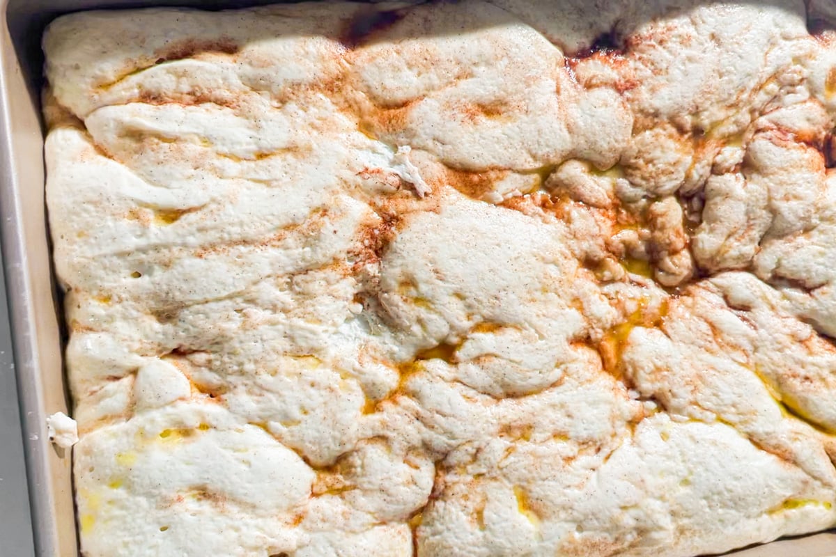 Close-up of a golden-brown, fluffy baked surface with swirls of melted butter and patches of cinnamon, resembling a cinnamon roll in a rectangular baking pan.
