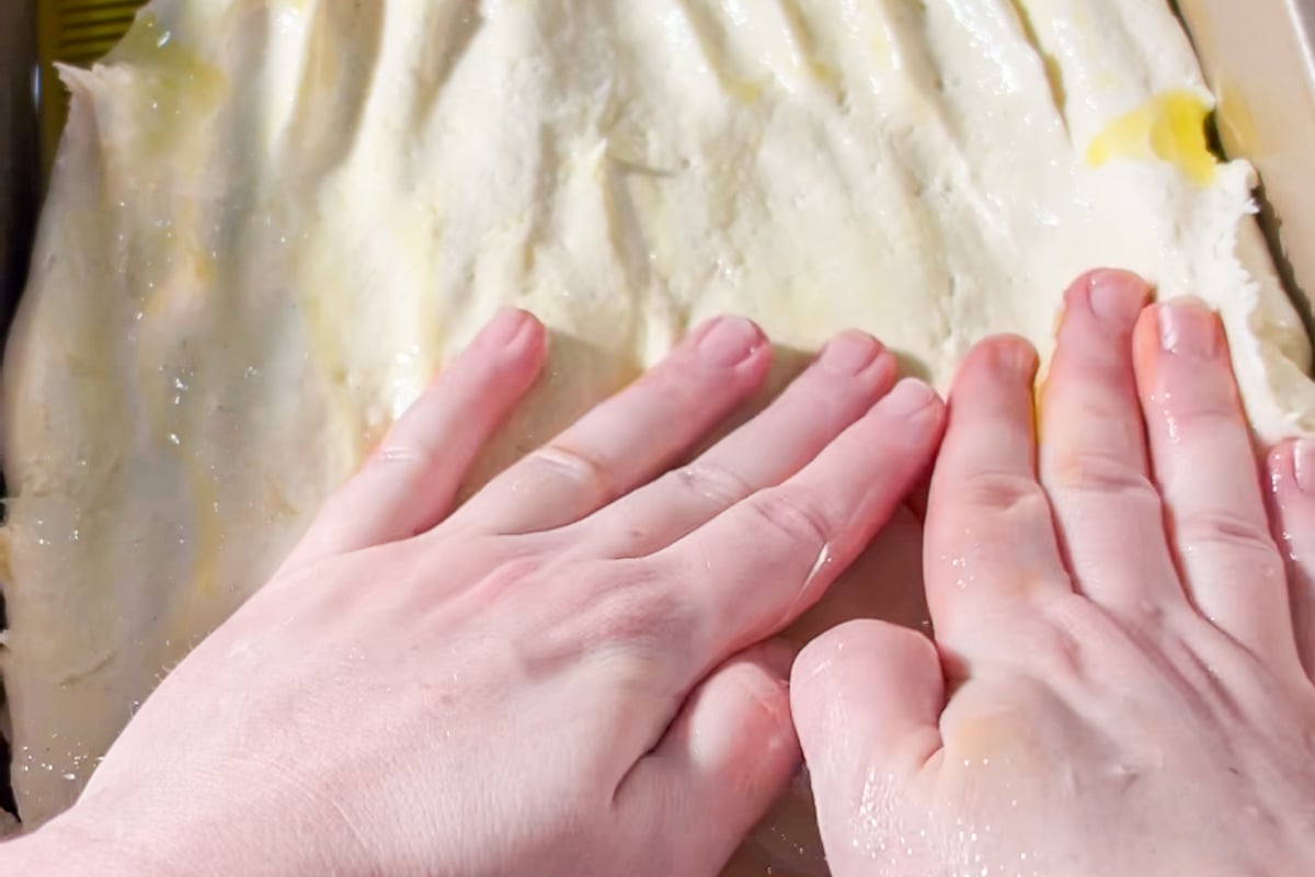 Two hands pressing and spreading dough in a rectangular baking pan, with a light layer of oil visible on the dough’s surface.