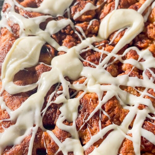 Close-up of a cinnamon pull-apart focaccia topped with drizzles of white icing, sitting on a cooling rack. The bread has a golden-brown, textured surface with visible cinnamon swirls.