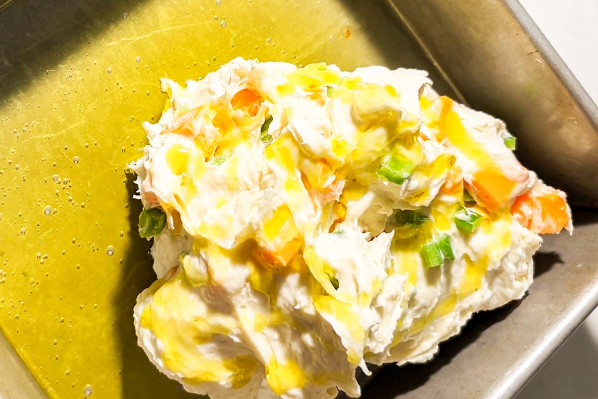 A close-up of a metal baking pan with a ball of gluten-free focaccia dough containing chopped jalapeños and cubed cheddar cheese, drizzled with olive oil.