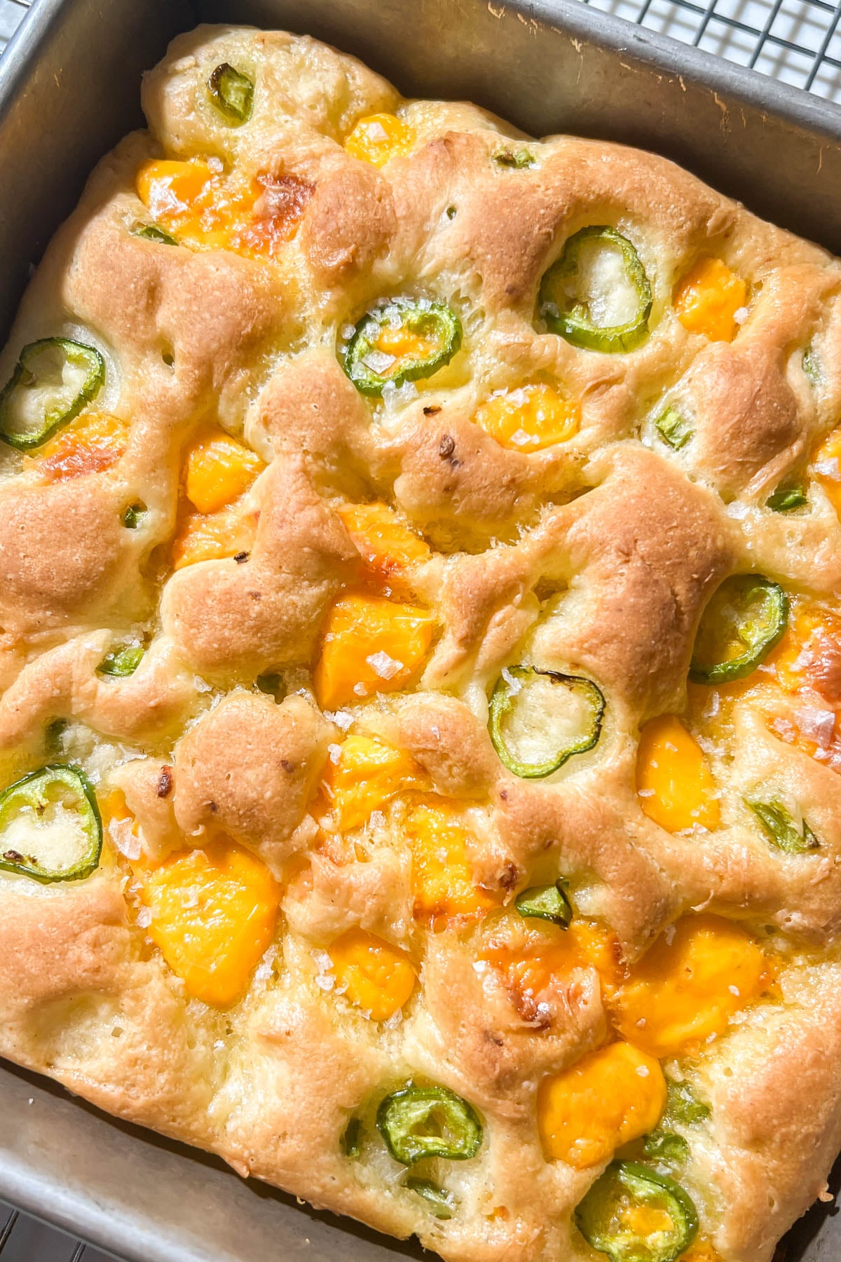 Golden brown focaccia bread topped with melted cheddar cheese and sliced jalapeños, shown in a baking pan on a wire rack. The bread appears soft and fluffy with visible air pockets.