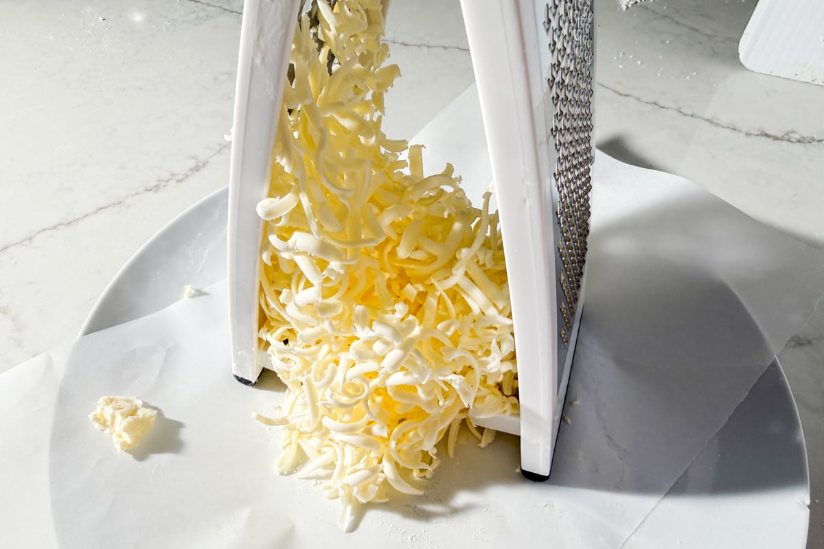A close-up of shredded cheese falling from a box grater onto a white plate on a marble countertop.