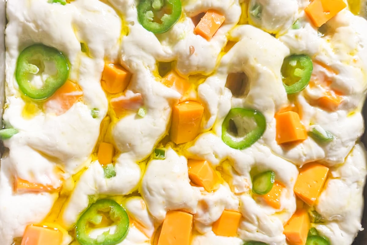 Close-up of raw focaccia dough topped with sliced jalapeños, cubes of cheddar cheese, and drizzled with olive oil, ready to be baked.