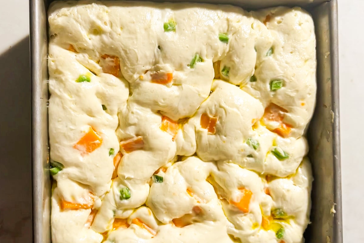 Unbaked gluten-free focaccia dough in a square pan, with visible chunks of cheddar cheese and diced jalapeños mixed throughout the dough, ready to be baked.