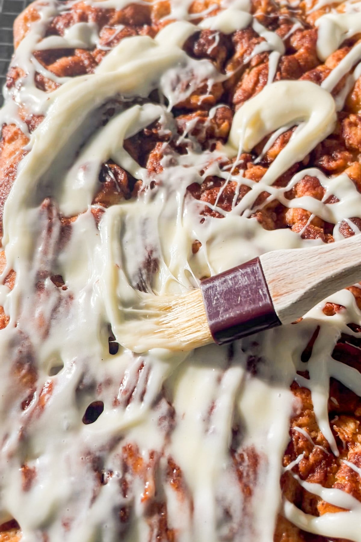 A close-up of gluten-free cinnamon roll focaccia being topped with cream cheese frosting. A pastry brush is spreading the icing across the surface, coating the golden-brown dough beneath.