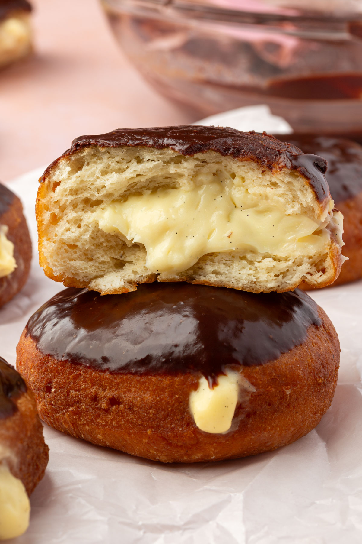 A stack of two chocolate-glazed, cream-filled donuts sits on parchment paper. The top donut is sliced open, revealing a rich vanilla custard filling oozing out.
