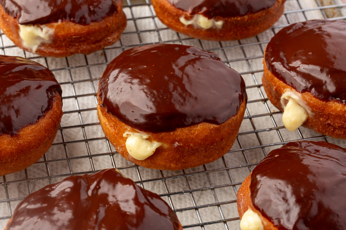 Close-up of several Boston cream donuts on a wire cooling rack. The donuts are topped with shiny chocolate glaze and filled with creamy custard, some of which is visible where it oozes out from the sides.