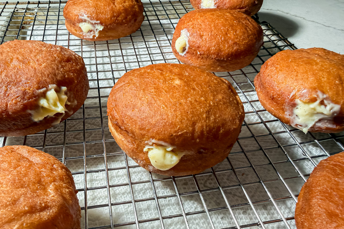 Golden brown cream-filled donuts rest on a wire cooling rack, with some cream visibly oozing from the sides, set on a light-colored surface.