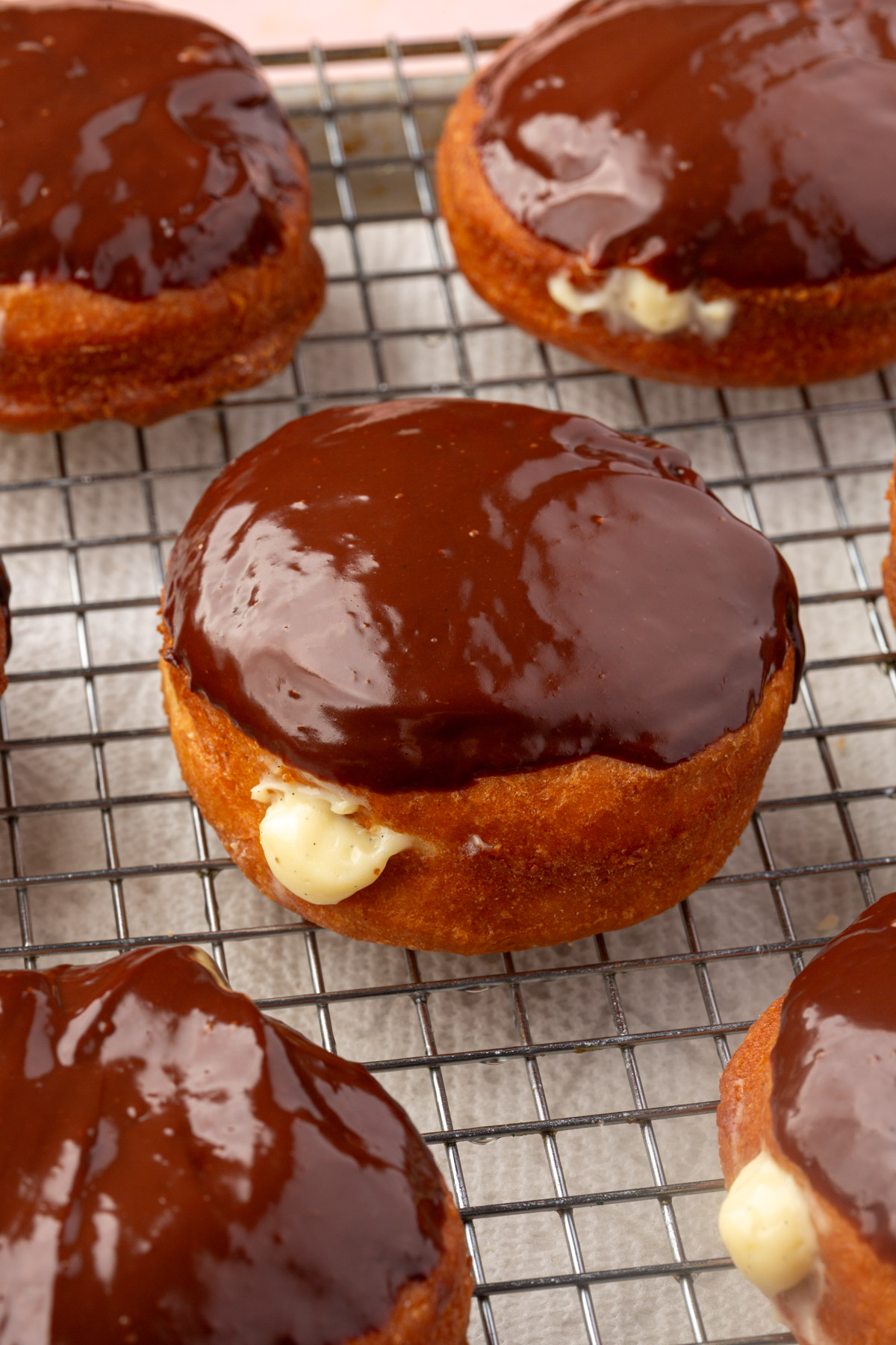 Boston cream donuts with chocolate glaze and creamy filling are cooling on a wire rack lined with parchment paper.