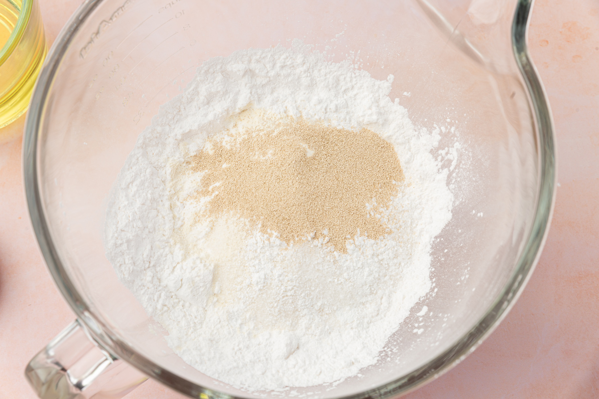 A glass mixing bowl containing white flour and a small mound of dry yeast on a light pink surface.