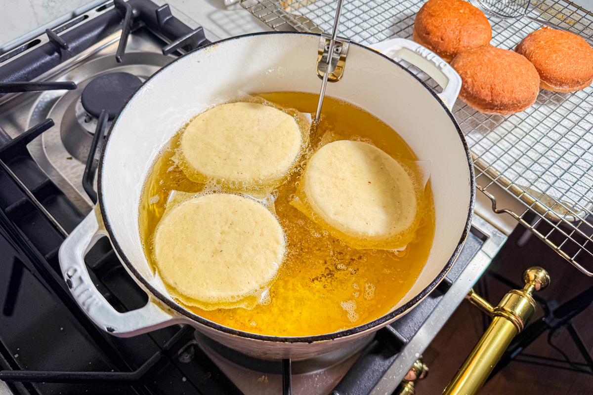 Three round pieces of dough are being deep-fried in a pot of oil on a stove. Next to the pot, fried doughnuts are cooling on a wire rack.