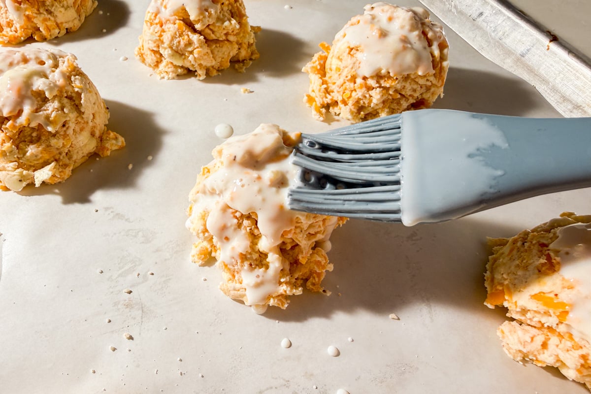 A silicone brush applies a white glaze to unbaked biscuit dough mounds on a parchment-lined baking sheet.