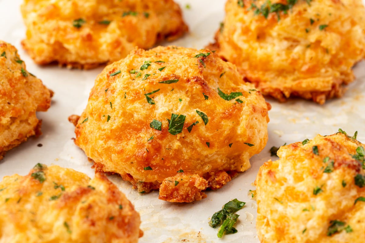 Close-up of golden brown, cheesy biscuits sprinkled with chopped parsley, resting on a sheet of parchment paper. The biscuits look fluffy and slightly crispy on the edges.