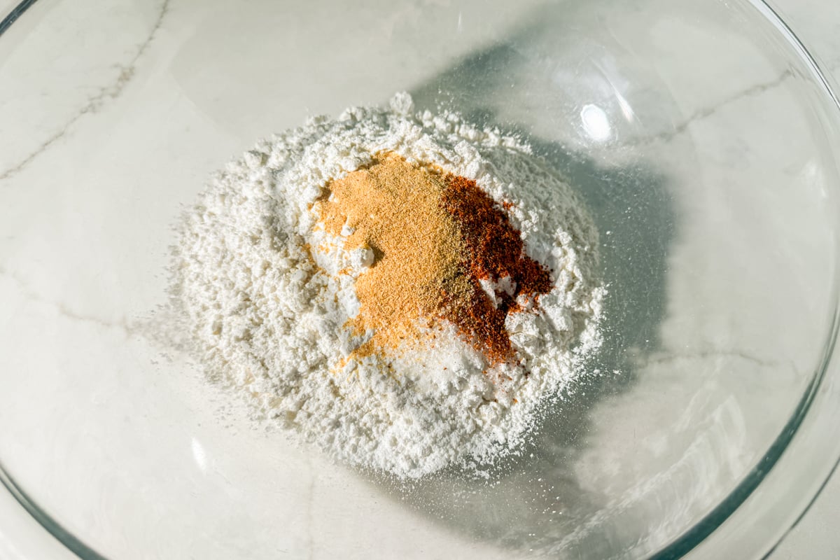 A glass bowl containing a mixture of white flour and various ground spices, including yellow and red powders, on a light-colored countertop.
