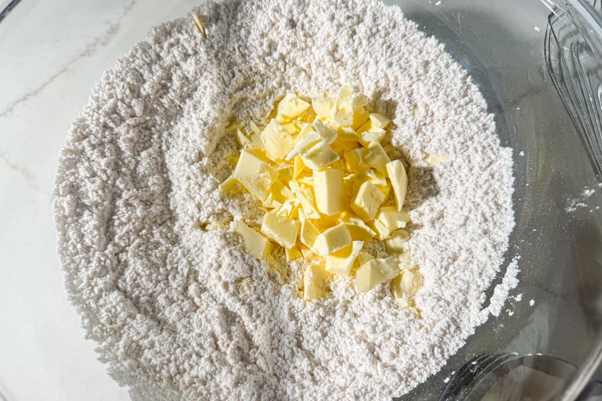 A glass bowl filled with flour and small chunks of butter, ready to be mixed, sits on a light countertop.