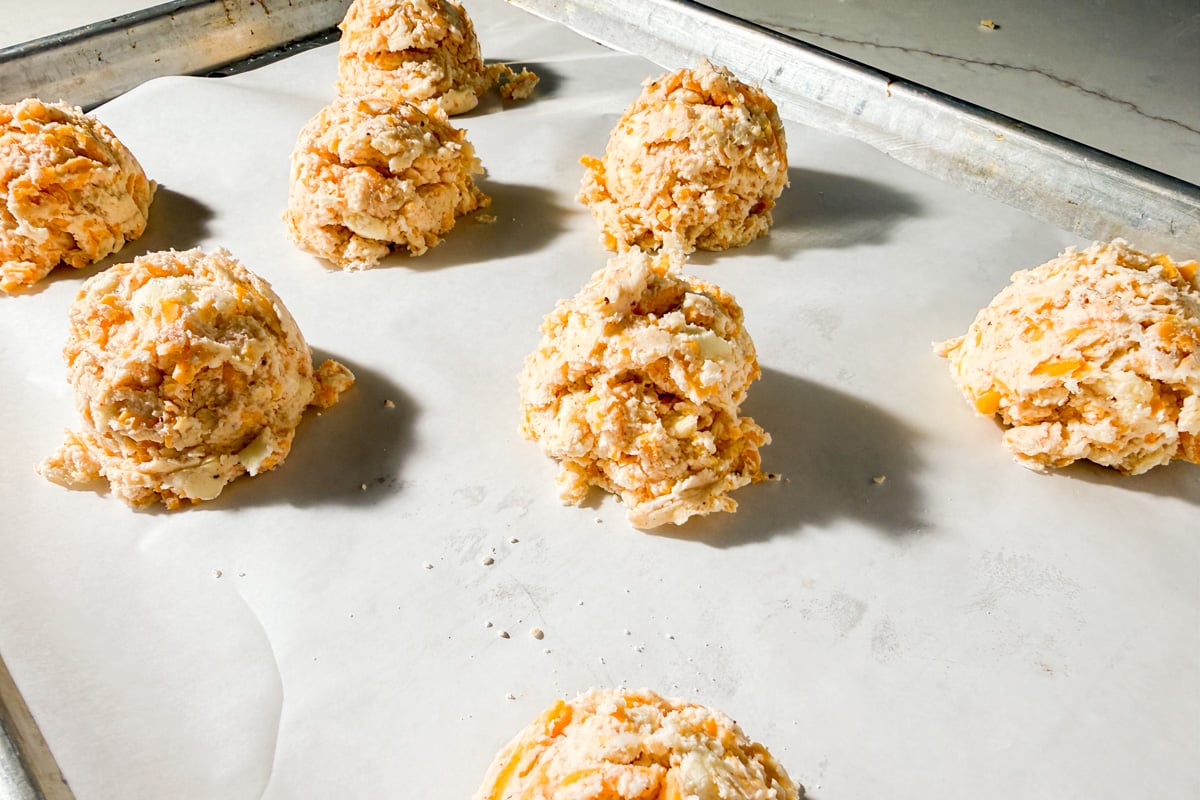 Scoops of unbaked biscuit dough with visible cheese pieces sit spaced apart on a parchment-lined baking sheet, ready to be baked.