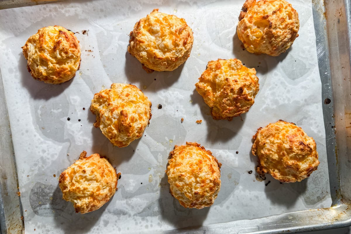 Golden, freshly baked drop biscuits are spread out on a parchment-lined baking sheet, with a few browned spots indicating crisp edges. The biscuits appear fluffy and slightly uneven in shape.