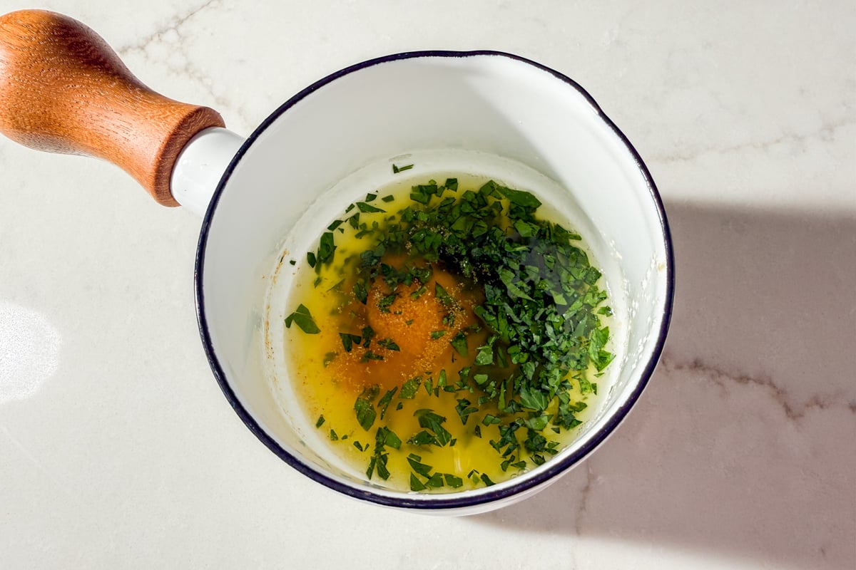 A white saucepan with a wooden handle contains melted butter, chopped fresh herbs, and spices, placed on a white marble surface.