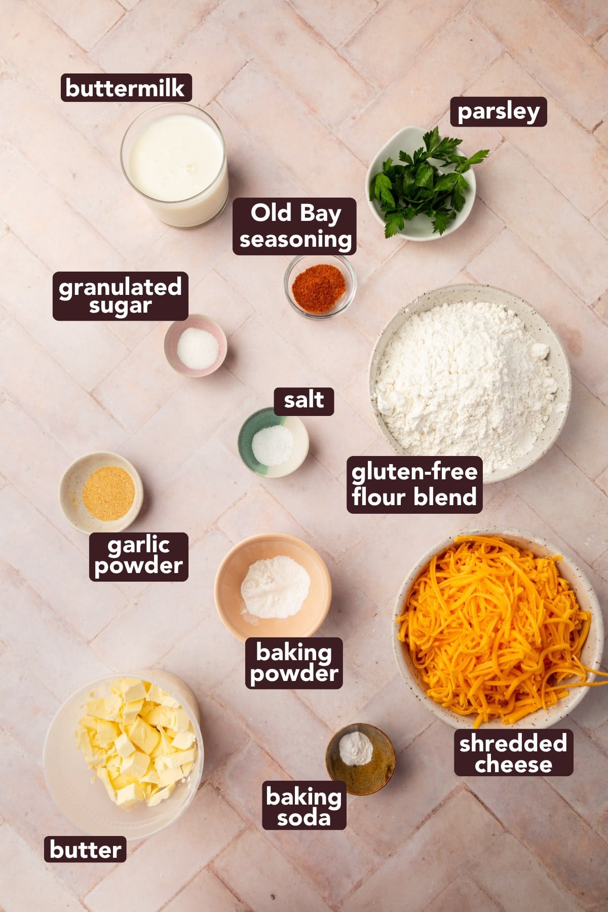 Top-down view of labeled ingredients in bowls and cups, including buttermilk, granulated sugar, Old Bay seasoning, salt, parsley, gluten-free flour, garlic powder, baking powder, baking soda, shredded cheese, and butter.