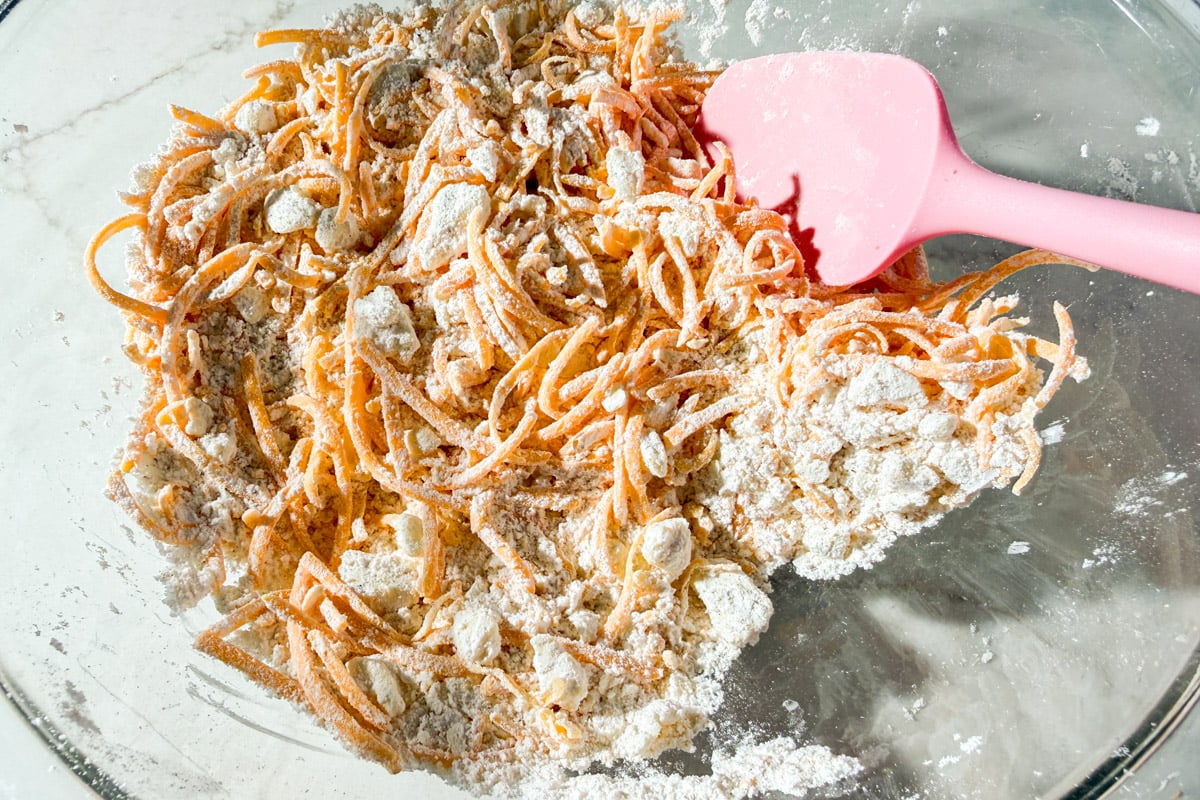 A glass bowl containing shredded carrots and flour, being mixed with a pink spatula. The mixture appears dry and partially combined, ready for the next baking step.