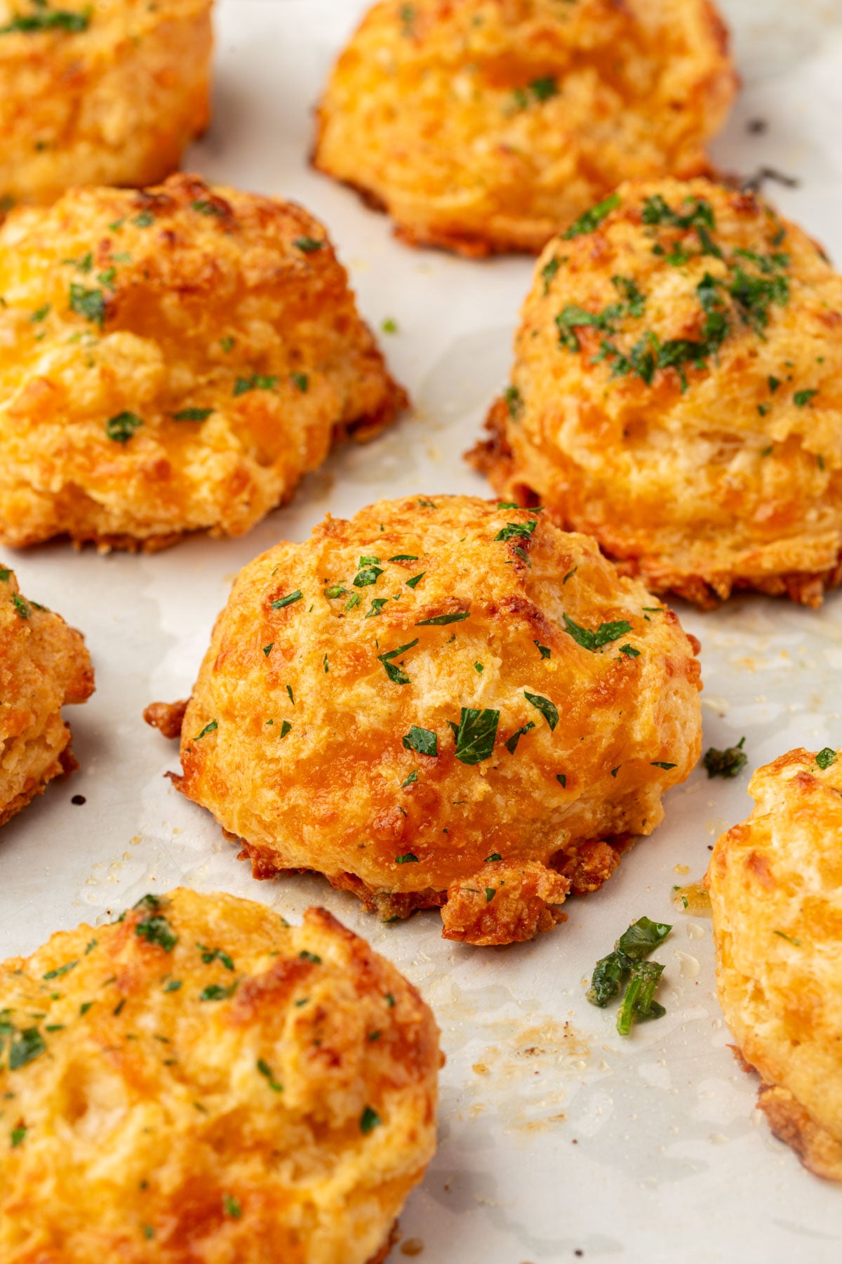 Golden, cheesy biscuits topped with chopped parsley are arranged on a sheet of parchment paper. The biscuits look freshly baked, with crispy edges and a soft, fluffy texture.