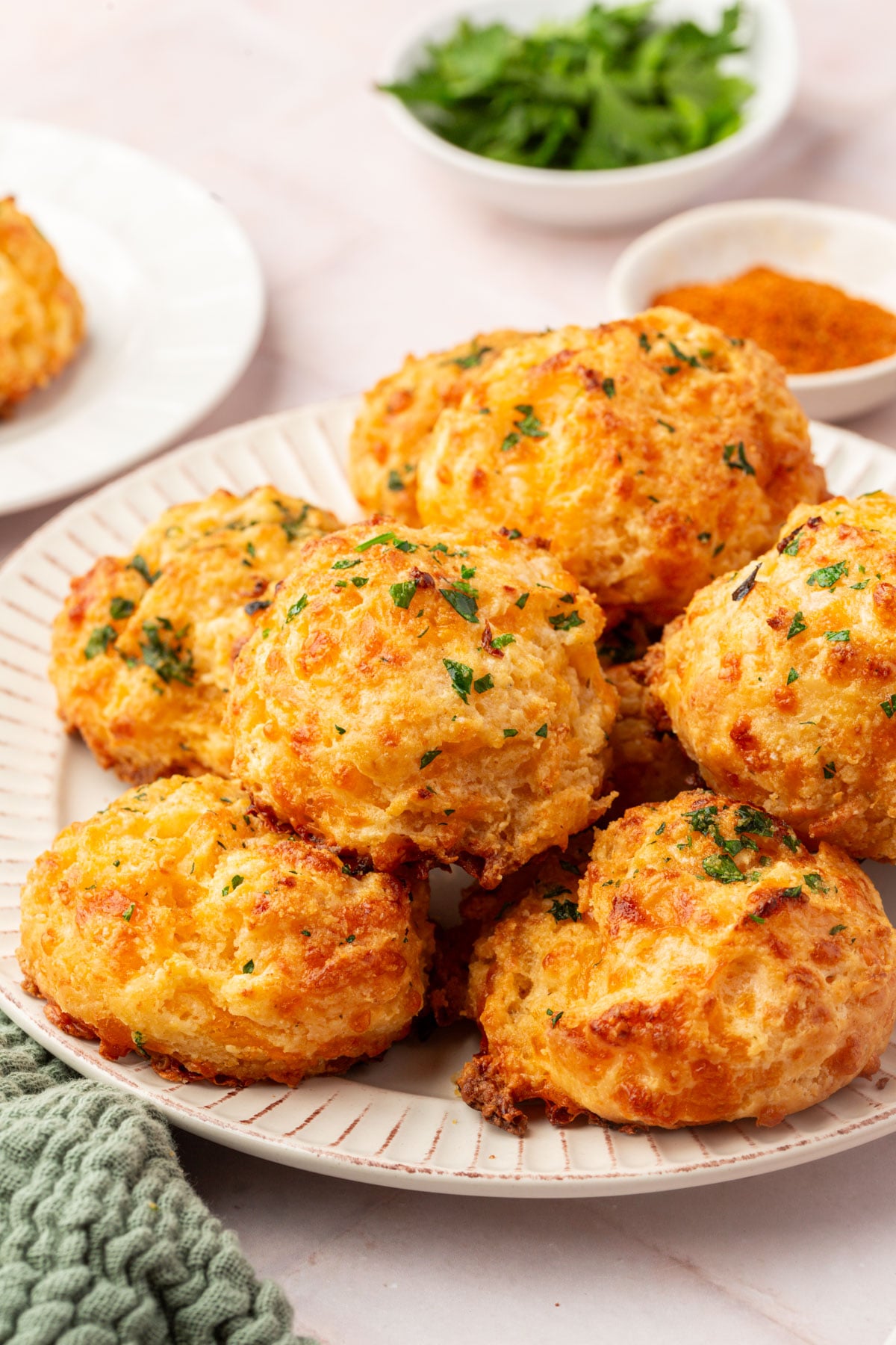 A plate piled with golden, cheesy biscuits garnished with chopped herbs. Small bowls of seasoning and fresh parsley, along with a green towel, are visible in the background on a light-colored table.