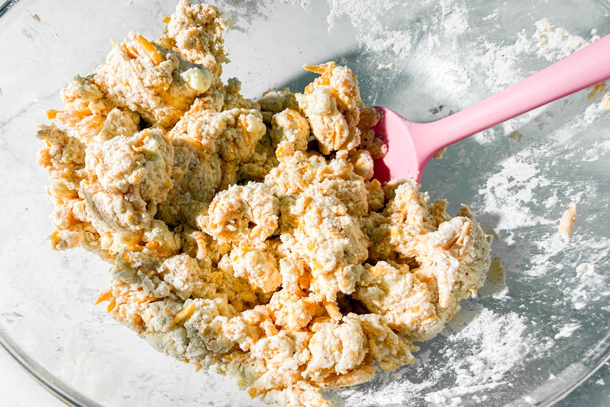 Rough, crumbly biscuit dough mixed with shredded cheddar cheese in a clear glass bowl, being stirred with a pink spatula.