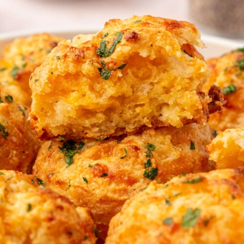 A close-up of golden, cheesy biscuits garnished with chopped parsley, with one biscuit broken open to show a soft, fluffy interior. A blurred bowl of herbs appears in the background.
