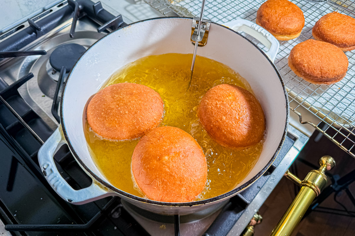 Three round doughnuts are frying in hot oil inside a white pot on a stove, with a metal thermometer clipped to the pot. Finished doughnuts cool on a wire rack nearby.