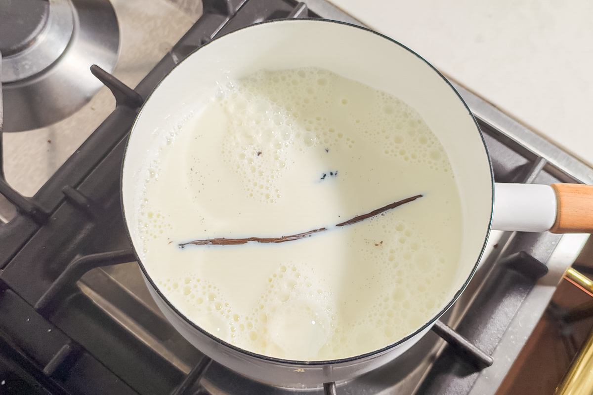 A white saucepan with milk and a split vanilla bean heating on a stovetop burner. Bubbles are forming on the surface of the milk.