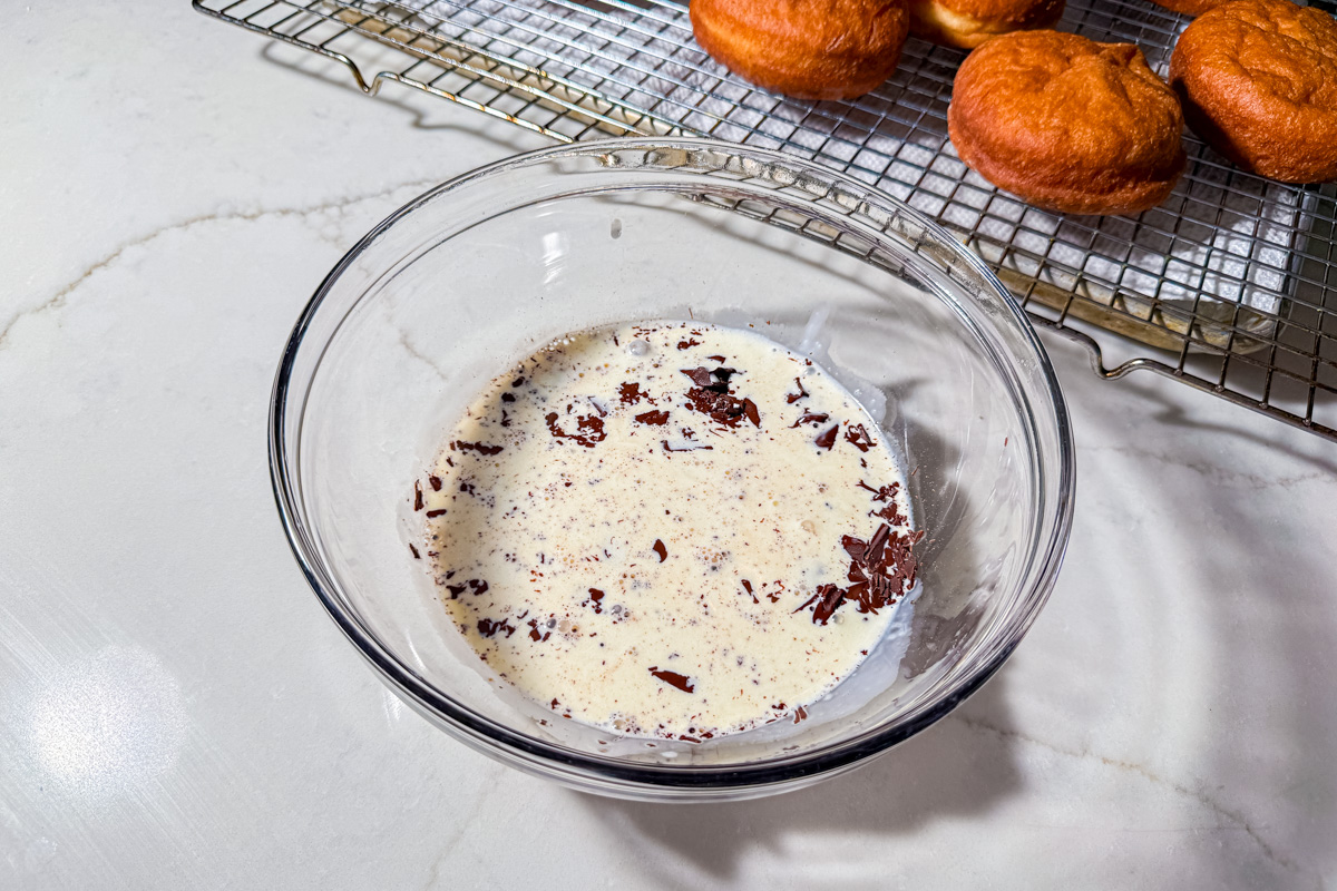 A glass bowl with cream and chopped chocolate sits on a white countertop. In the background, fried pastries cool on a metal wire rack.