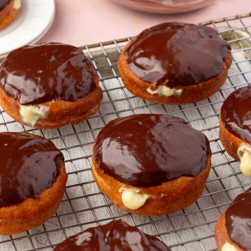 Boston cream donuts with shiny chocolate glaze and creamy filling are arranged on a wire cooling rack, with a bowl of chocolate glaze and a plate in the background.