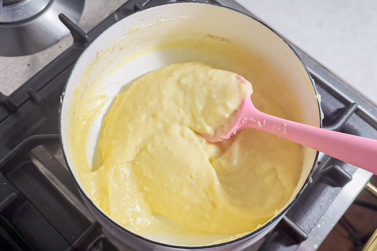 A pink spatula stirs thick, yellow pastry cream in a white saucepan on a stovetop.
